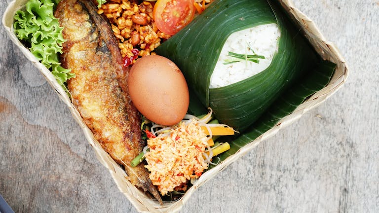 Close-up of traditional Asian cuisine featuring rice, fish, egg, and vegetables in a woven basket.